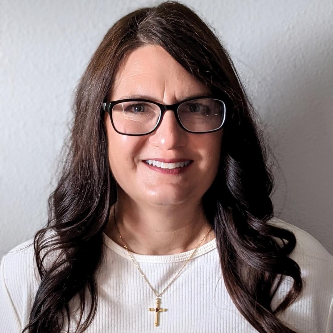 The woman in the foreground is wearing a white shirt, dark glasses with thick black rectangular frames, a gold cross on a ...